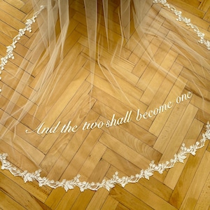 以下が含まれることがあります： 繊細な花のレースのトリムが付いた白いチュールのウェディングベール。ベールには「And the two shall become one」というテキストが刺繍されています。