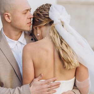 May include: A bride and groom embrace, the bride wearing a white strapless wedding dress with a tulle veil and a bow in her hair. The groom is wearing a beige suit jacket and a white shirt.