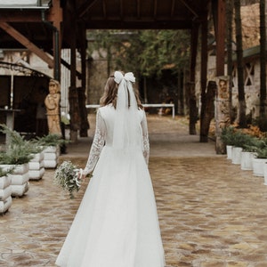 May include: A bride in a white wedding dress with a white bow in her hair walks down a stone path towards a wooden archway decorated with string lights.