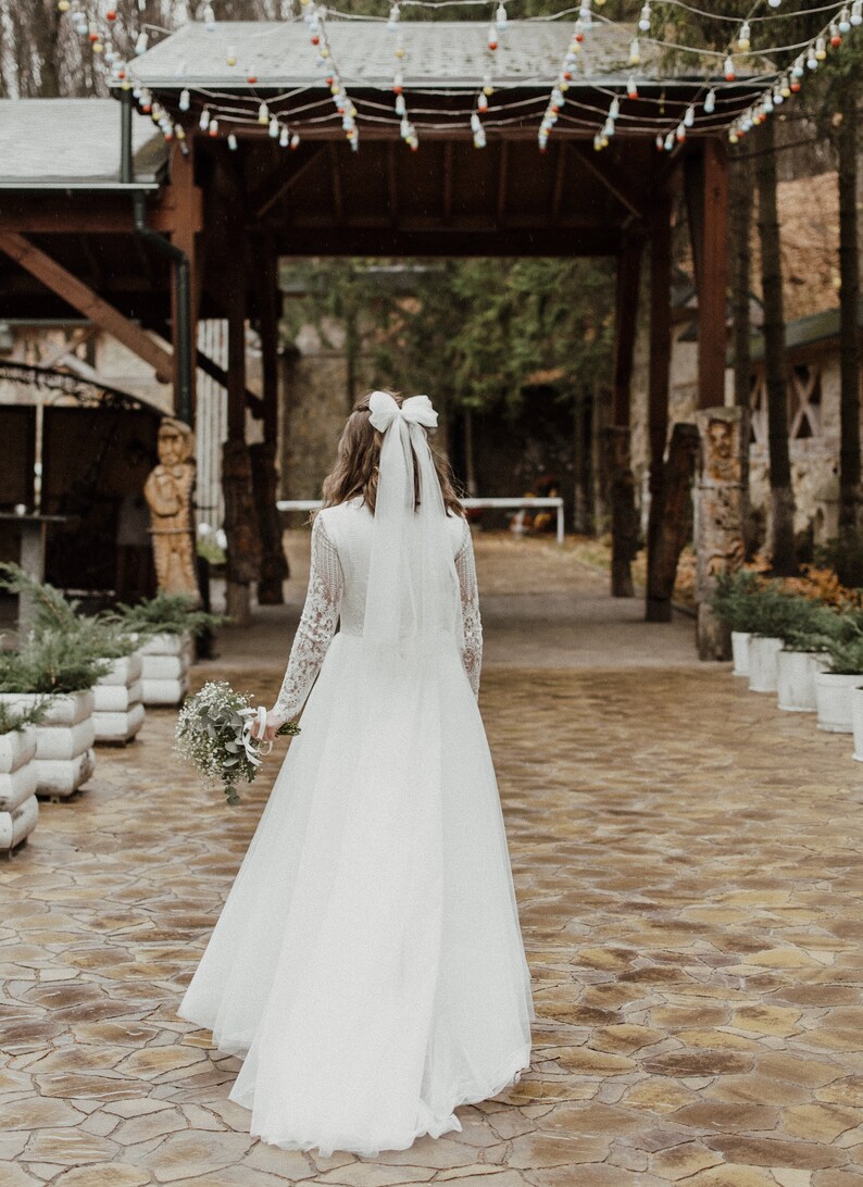 May include: A bride in a white wedding dress with a white bow in her hair walks down a stone path towards a wooden archway decorated with string lights.