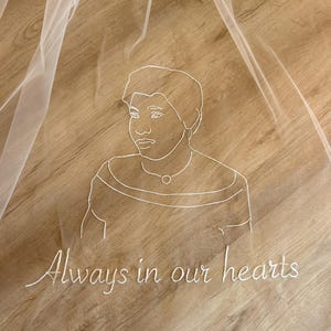 May include: A sheer white veil featuring a delicate white thread embroidery of a person's portrait in a continuous line style. Below the portrait, the text "Always in our hearts" is embroidered in a matching white cursive script font.