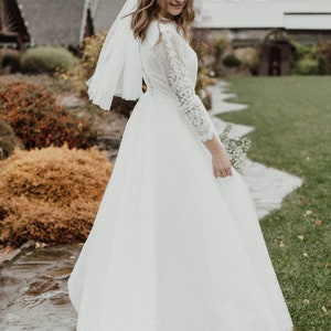 May include: A bride wearing a white lace wedding dress with a long veil and holding a bouquet of white flowers. She is standing on a path in front of a building.