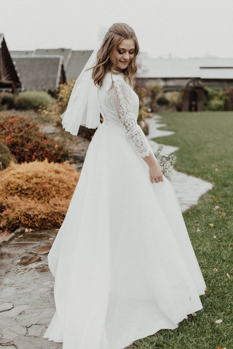 May include: A bride wearing a white lace wedding dress with a long veil and holding a bouquet of white flowers. She is standing on a path in front of a building.