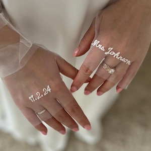 May include: Close-up of a bride's hands wearing rings and a sheer veil. The veil is embroidered with the text "Mrs. Johnson" and the date "11.2.24". The bride is wearing a wedding dress.