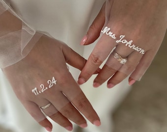 May include: Close-up of a bride's hands wearing rings and a sheer veil. The veil is embroidered with the text "Mrs. Johnson" and the date "11.2.24". The bride is wearing a wedding dress.