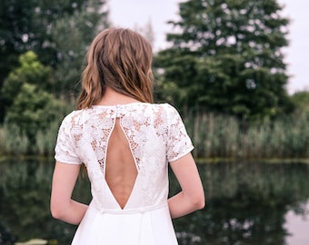 Può includere: Un abito da sposa bianco in pizzo con schiena scoperta e un'apertura a chiave. L'abito ha maniche corte e una gonna fluida.