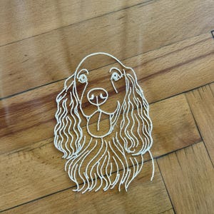 May include: White embroidered outline of a Cocker Spaniel dog's face. The dog's features include long, wavy ears, a smiling mouth, and detailed eyes. The embroidery is on a sheer, light-coloured fabric.