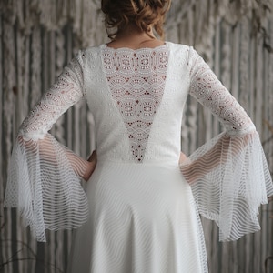 Pode incluir: Um vestido de noiva branco de renda com costas baixas e mangas sino. O vestido tem um corpete justo e uma saia fluida.