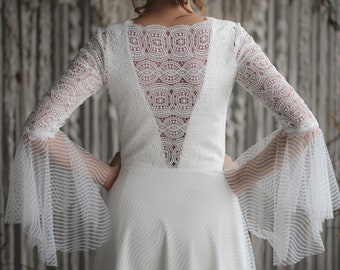 May include: A white lace wedding dress with a low back and bell sleeves. The dress has a fitted bodice and a flowing skirt.