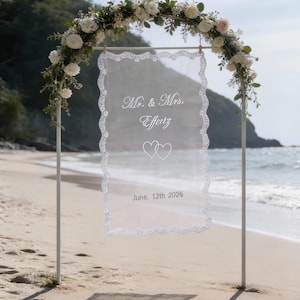 May include: A white wedding arch on a sandy beach, adorned with white flowers and greenery. A sheer, rectangular fabric panel hangs in the center, edged with lace and displaying the text "Mr. & Mrs. Effertz" and "June, 12th 2026".