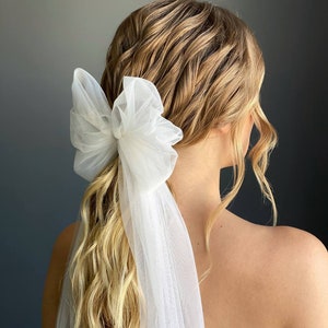 May include: A white tulle bow hair accessory with a long flowing veil. The bow is tied around a ponytail and the veil drapes down the back.