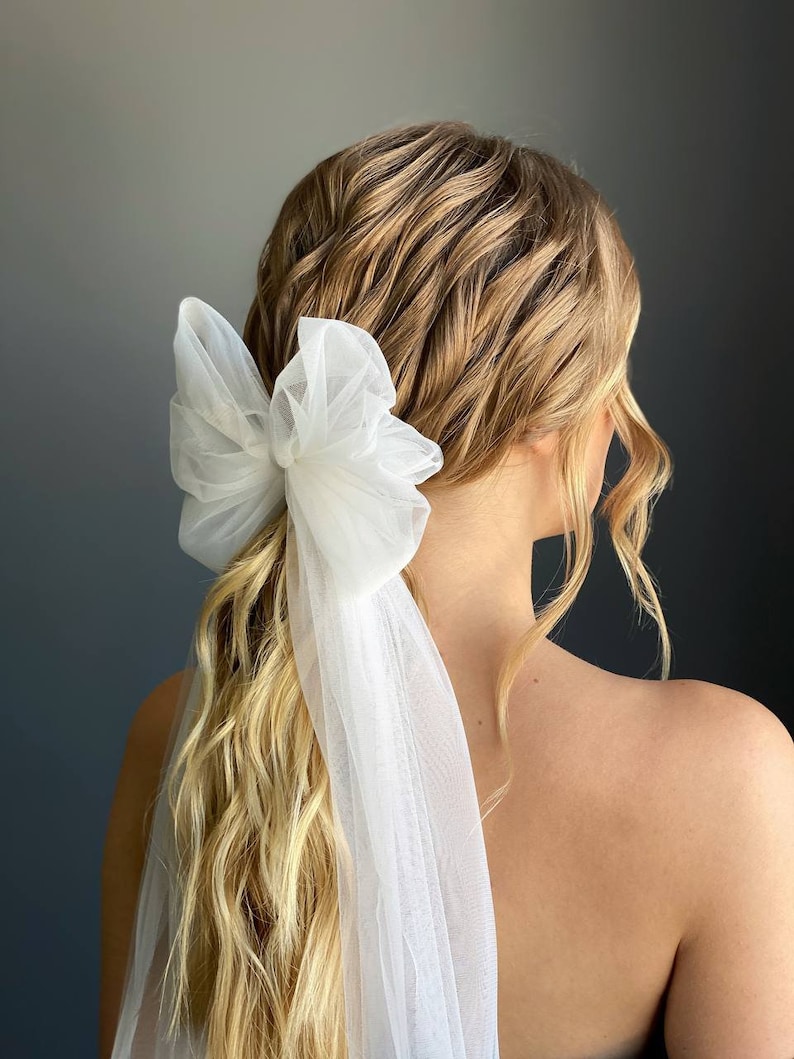 May include: A white tulle bow hair accessory with a long flowing veil. The bow is tied around a ponytail and the veil drapes down the back.