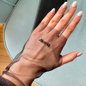May include: A sheer black glove with the word "NOW" printed on the palm. The glove is worn on a hand with long, oval-shaped, light-colored nails. A black object is in the background, and the hand rests on a light blue surface.