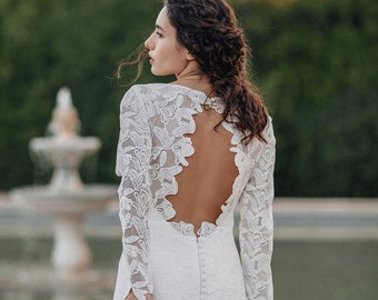 May include: A white lace wedding dress with a low back and long sleeves. The dress has a fitted bodice and a flowing skirt.