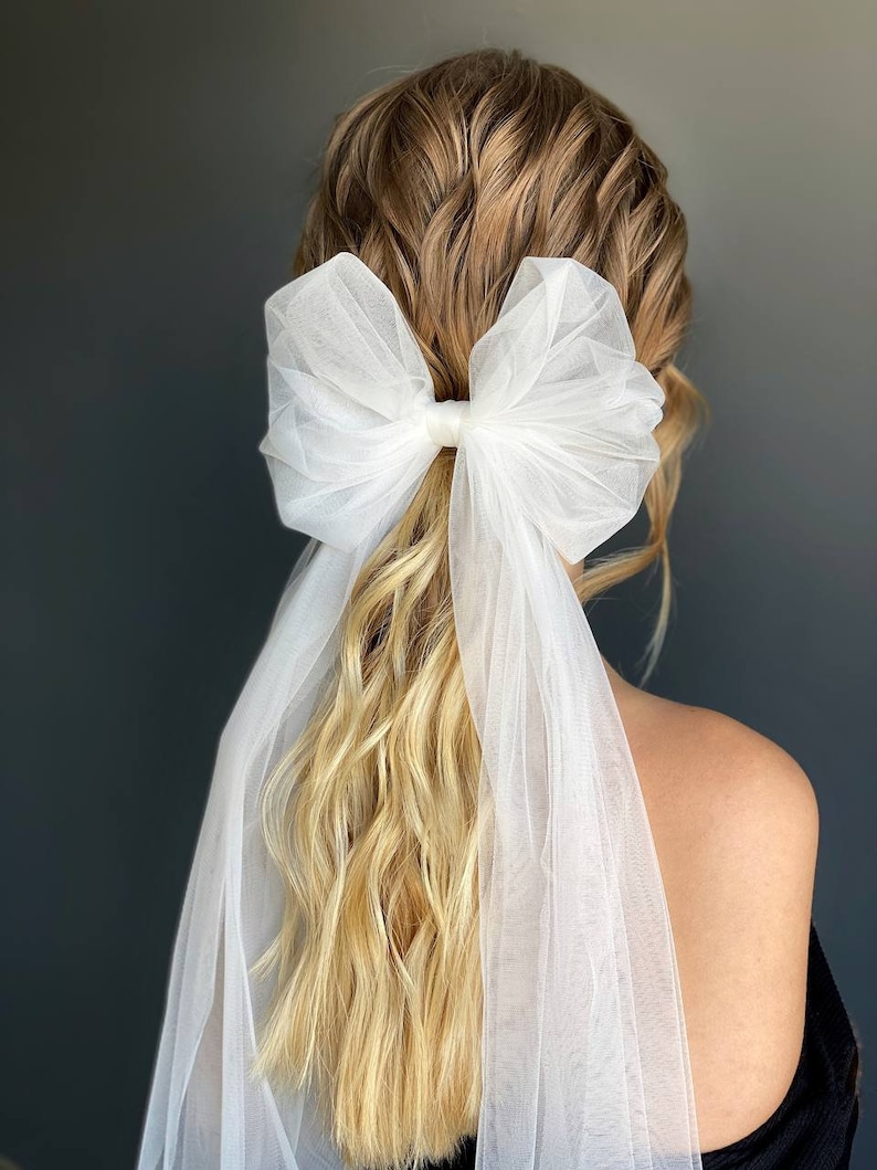 May include: A white tulle bow hair accessory with a long veil attached. The bow is tied in a large, loose knot and the veil drapes down the back of the head.