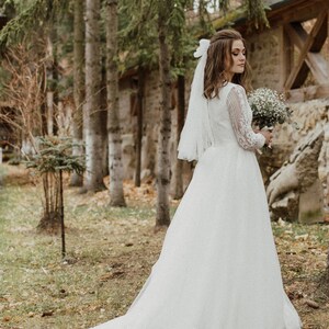 May include: A bride in a white lace wedding dress with a long veil and a bouquet of white flowers. She is standing in a forest setting.