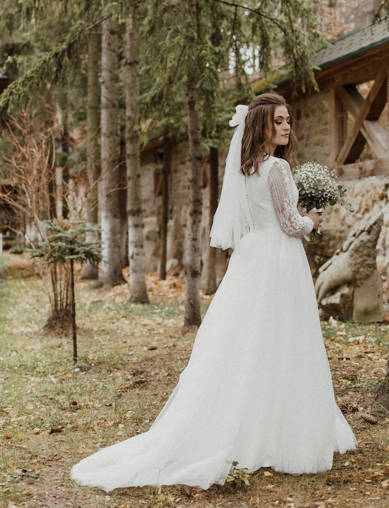May include: A bride in a white lace wedding dress with a long veil and a bouquet of white flowers. She is standing in a forest setting.