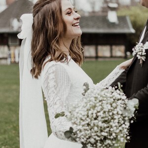May include: A bride in a white lace dress with a white veil and a bouquet of white baby's breath flowers smiles at the groom.