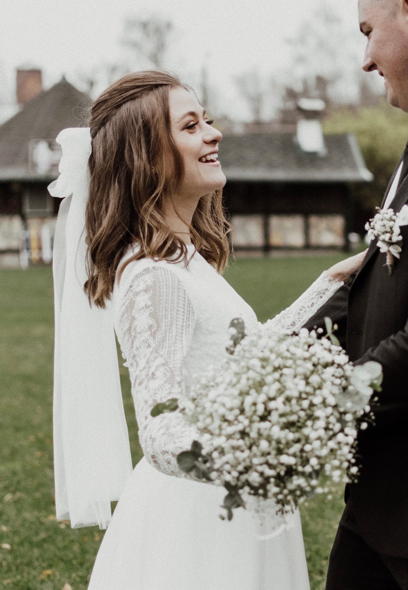 May include: A bride in a white lace dress with a white veil and a bouquet of white baby's breath flowers smiles at the groom.