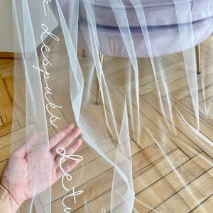 Puede incluir: Un largo velo de novia blanco y transparente hecho de tela de tul fina. El velo presenta una elegante inscripción blanca en cursiva bordada a lo largo del borde, mostrando la frase "amor después de tu amor" en un estilo de letra fluido.