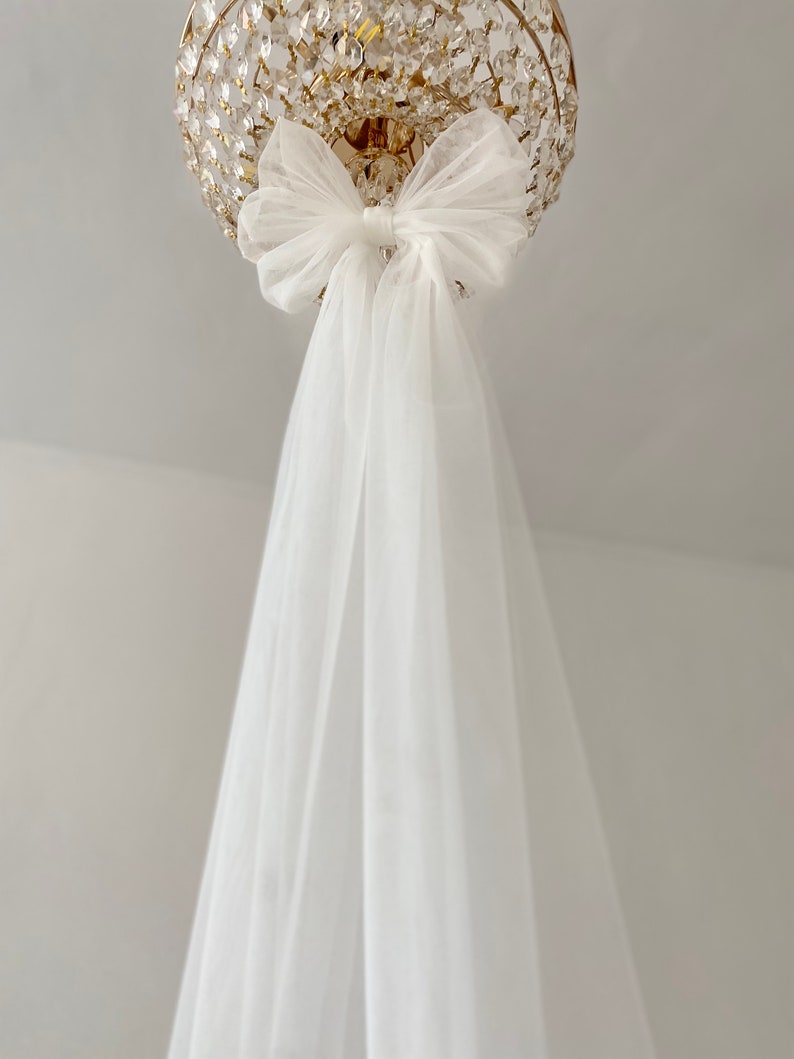 May include: A white tulle bow tied around a gold chandelier. The tulle drapes down from the bow.