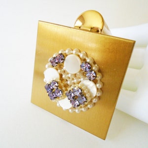 May include: A gold-coloured square compact with a decorative circular design. The design features faux pearls, white accents, and purple gemstones. The compact has a gold clasp.