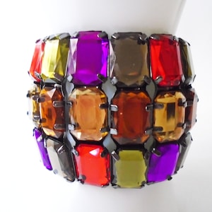 May include: A multi-colored bracelet with large, faceted, rectangular gems set in a black metal frame. The gems are in shades of red, orange, yellow, green, and purple.