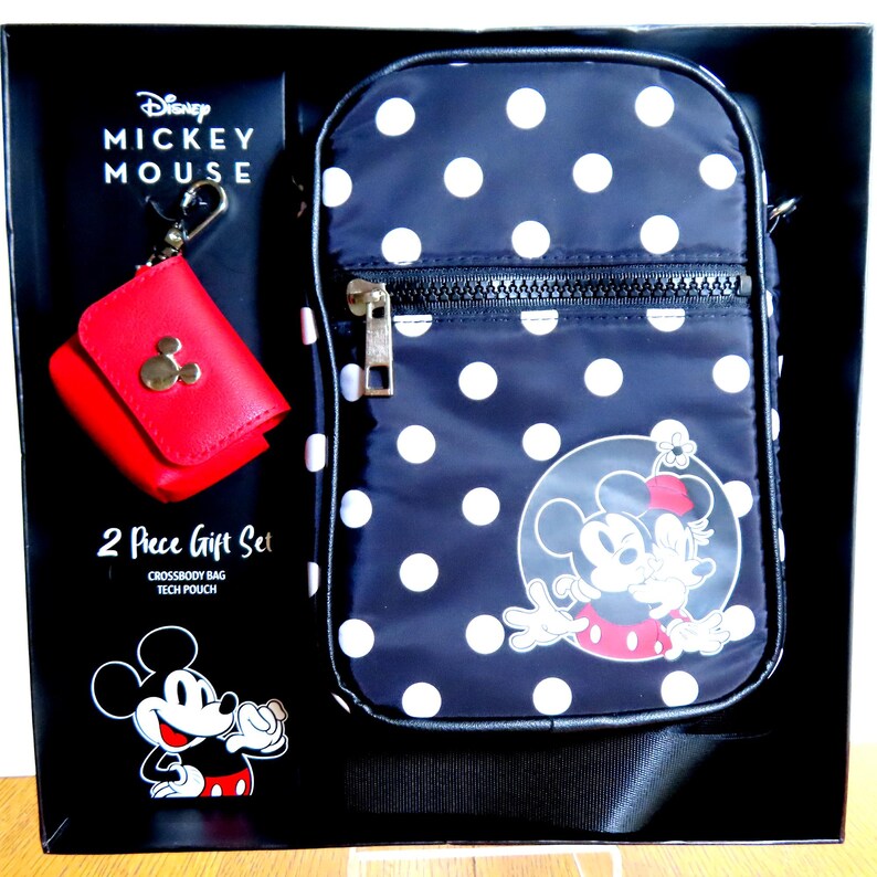 May include: A two-piece gift set featuring a navy blue crossbody bag with white polka dots and a red tech pouch. The bag has a Mickey and Minnie Mouse graphic. The set is packaged in a black box with Disney branding.