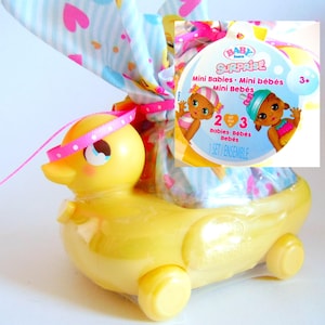 May include: A yellow plastic duck-shaped container with a pink and white polka-dot ribbon and a bag of toys. The bag has a blue and white pattern with hearts and a tag that says "BABY born SURPRISE Mini Babies".