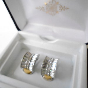 May include: A pair of silver earrings with clear crystal stones and a gold accent. The earrings are in a white box with a gold design.