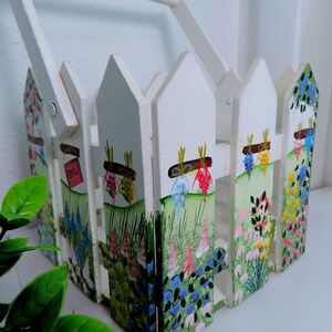 May include: A white wooden basket shaped like a picket fence, featuring hand-painted floral designs with green grass, colorful flowers, and small hanging bundles. The basket includes a sturdy white handle attached to the sides with metal hardware.