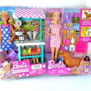 May include: Two Barbie doll sets. One features a doll in a blue floral dress with a market stand, and the other includes a doll in a pink checkered dress with a dog and accessories. Both sets are in their original packaging.