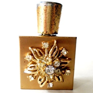 May include: A gold metal perfume bottle with a square base and a gold metal cap. The bottle is decorated with a gold metal flower design with clear rhinestones.
