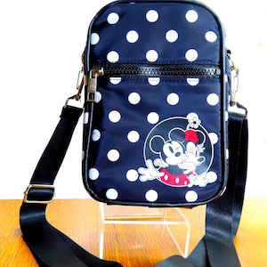 May include: Navy blue crossbody bag with white polka dots. Features a graphic of Minnie Mouse and Mickey Mouse. The bag has a black adjustable strap and a zipper closure.