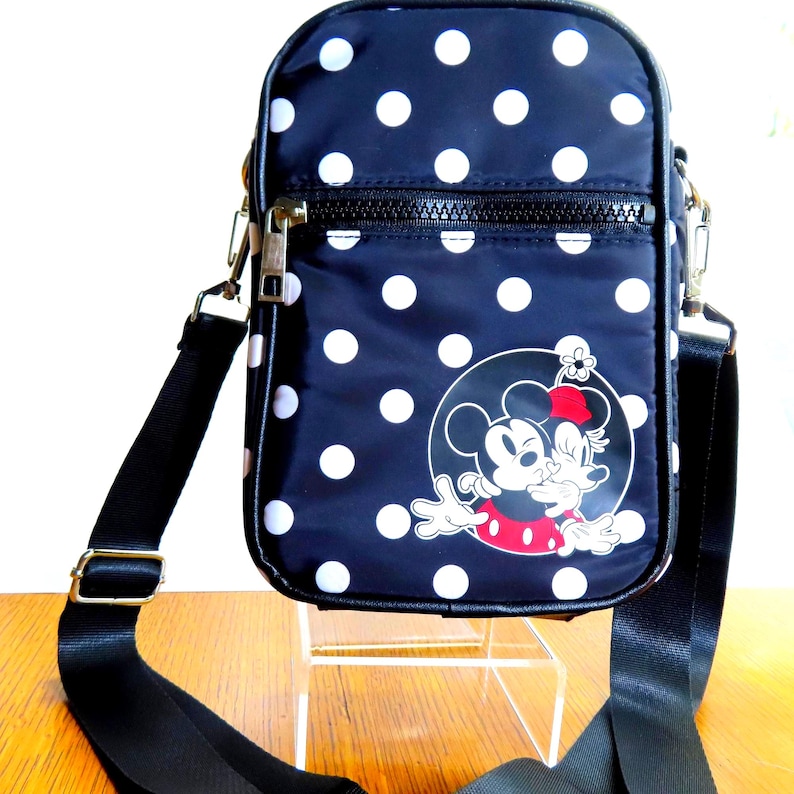 May include: Navy blue crossbody bag with white polka dots. Features a graphic of Minnie Mouse and Mickey Mouse. The bag has a black adjustable strap and a zipper closure.