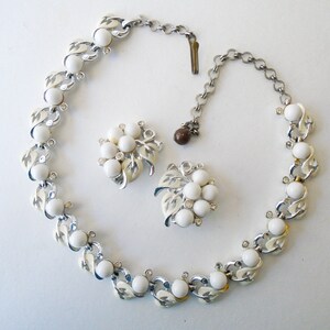 Op de afbeelding: Een witte en zilveren ketting en oorbellenset met een bladontwerp en witte ronde stenen. De ketting heeft een zilveren ketting met een sluiting.