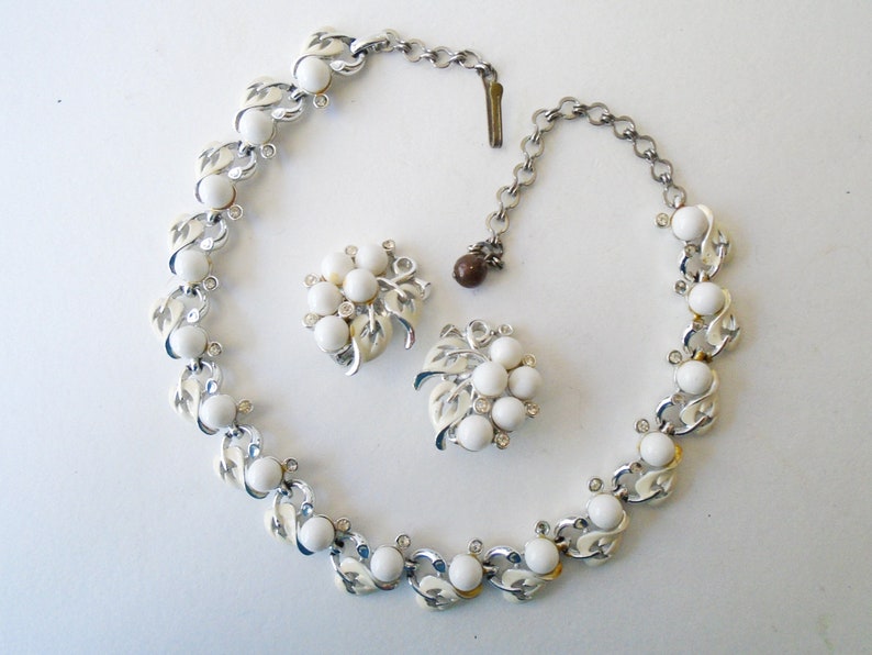 Op de afbeelding: Een witte en zilveren ketting en oorbellenset met een bladontwerp en witte ronde stenen. De ketting heeft een zilveren ketting met een sluiting.