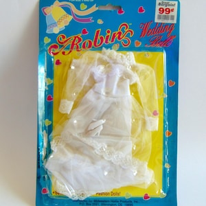 May include: A vintage doll wedding dress set in a plastic package. The white dress has lace trim and a sheer veil. The packaging is blue and yellow with the text "Robin's Wedding Belle" and "Great Bargain! 99¢".