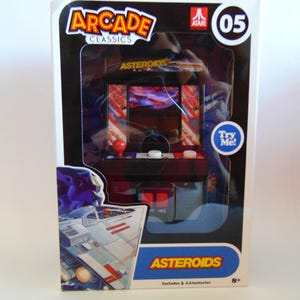 May include: Miniature "Asteroids" arcade game in a clear plastic box. The box features the "Arcade Classics" logo and the Atari logo. The game has a red joystick and white buttons. The box also says "Try Me!" and "Includes 3 AA batteries".