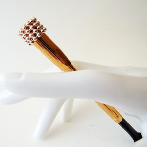 May include: A gold pen with a silver and white beaded top. The pen is resting on a white hand.