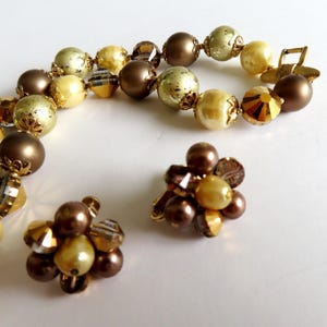 May include: A vintage jewelry set featuring a necklace and matching earrings. The necklace has large, round beads in shades of brown, gold, and cream. The earrings are clusters of smaller beads in similar colors. The set is displayed on a white surface.