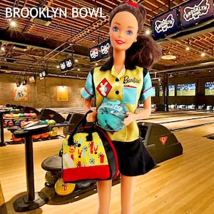 May include: A Barbie doll in a bowling alley setting. The doll wears a yellow and blue bowling shirt and black shorts, holding a bowling ball and a bowling bag. The bag is yellow, red, and black with a colorful design. The text "BROOKLYN BOWL" is visible.