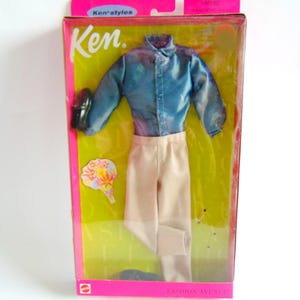 May include: A Ken doll fashion set in a pink and yellow box. The set includes a blue button-down shirt, tan pants, black shoes, and a floral accessory. The packaging reads "Ken" and "Fashion Avenue".
