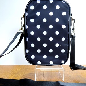 May include: Black crossbody bag with white polka dots. The bag has a black strap and silver-colored hardware. The bag is rectangular with rounded corners and a black trim. The bag is sitting on a clear stand.