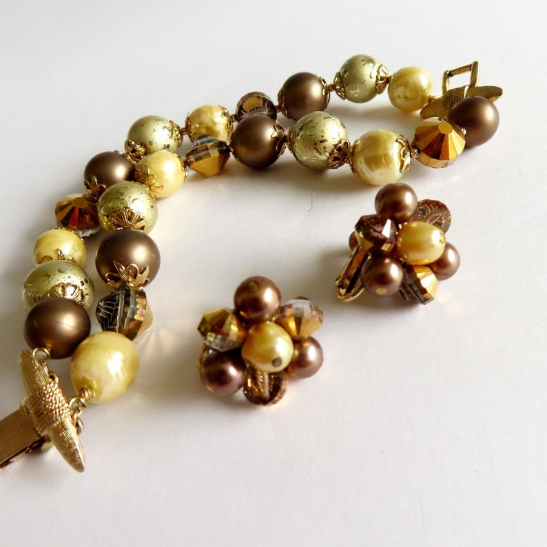 May include: A vintage jewelry set featuring a necklace and two pairs of clip-on earrings. The necklace and earrings are adorned with a mix of round and faceted beads in shades of gold, brown, and cream. The necklace has a gold-tone clasp.
