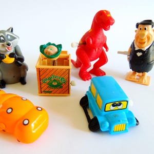 May include: A collection of vintage wind-up toys, including a raccoon, a red dinosaur, a blue car, a Fred Flintstone figure, a yellow hippo, and a Cabbage Patch Kids jack-in-the-box. The toys are made of plastic and are set against a white background.