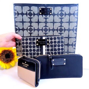 May include: A selection of Kate Spade wallets and a tote bag. The tote bag features a black and white spade design. The wallets are black, tan, and white with gold zips and hardware. A yellow sunflower is in the background.