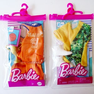 May include: Two Barbie doll clothing sets in clear plastic bags with pink hangers. The set on the left includes an orange basketball jersey with the text "MALIBU 6" on the front, orange shorts, a gold trophy, and a basketball. The set on the right includes a yellow short-sleeved shirt, green camouflage pants, and white sneakers. Both bags have the Barbie logo and the text "Ken" on the top.