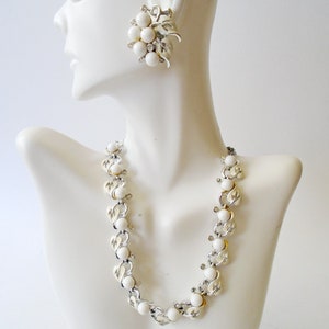 Op de afbeelding: Een witte en zilveren ketting en oorbellenset met grote witte kralen. De ketting is een ketting met een herhalend patroon van witte kralen en zilveren schakels. De oorbellen zijn een cluster van witte kralen met zilveren accenten.