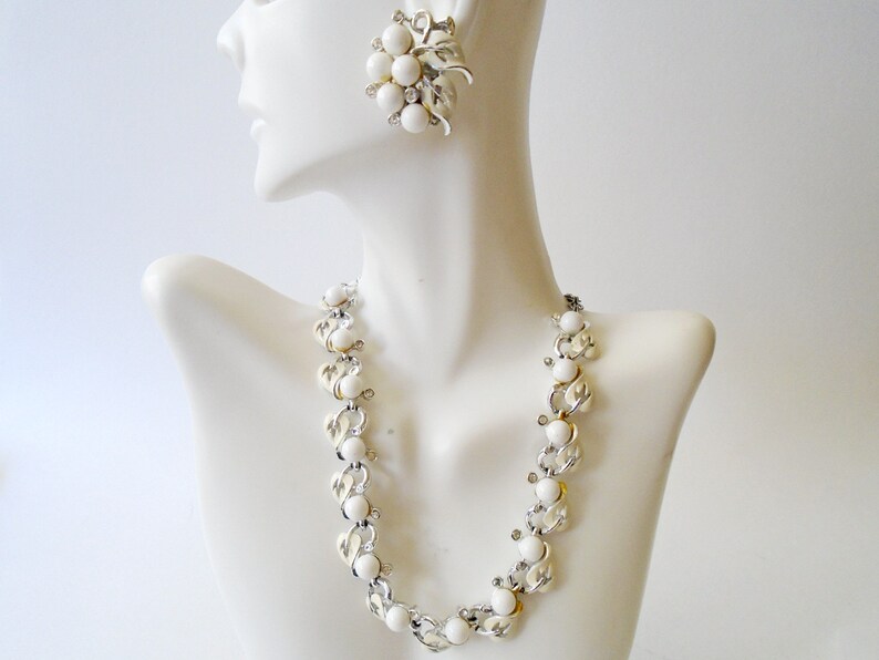 Op de afbeelding: Een witte en zilveren ketting en oorbellenset met grote witte kralen. De ketting is een ketting met een herhalend patroon van witte kralen en zilveren schakels. De oorbellen zijn een cluster van witte kralen met zilveren accenten.